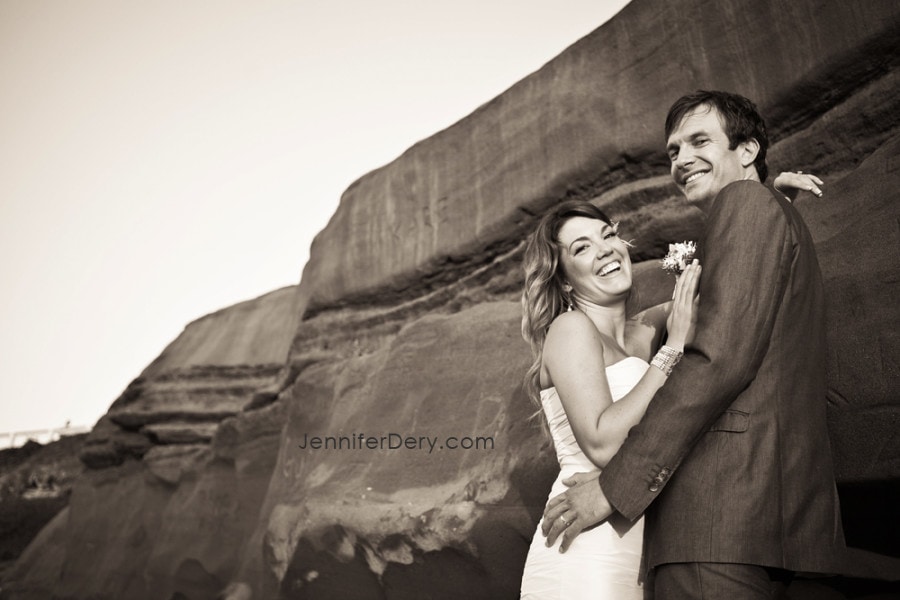 A couple in formal attire embraces and smiles joyfully by a rocky cliff. The woman wears a strapless dress, and the man is in a suit. The background features layered rock formations.