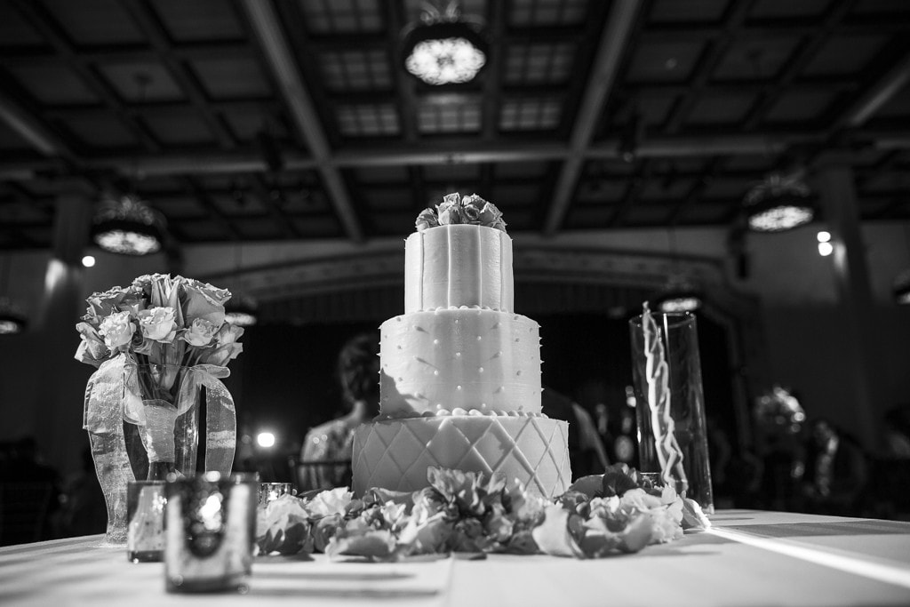 Black and white photo of a two-tiered wedding cake on a table decorated with flowers and candles. The background features ornate ceiling lights in a banquet hall setting.