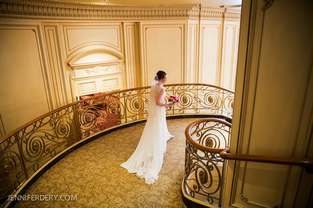 A bride in a white gown holds a bouquet of pink flowers as she stands on an ornate, curved staircase. The background features elegant, cream-colored paneling and a patterned carpet.