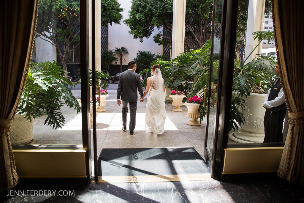 A bride and groom hold hands and walk through a doorway, surrounded by lush greenery and large planters with pink flowers. Sunlight filters through the trees onto the pavement outside, creating a warm and inviting setting.