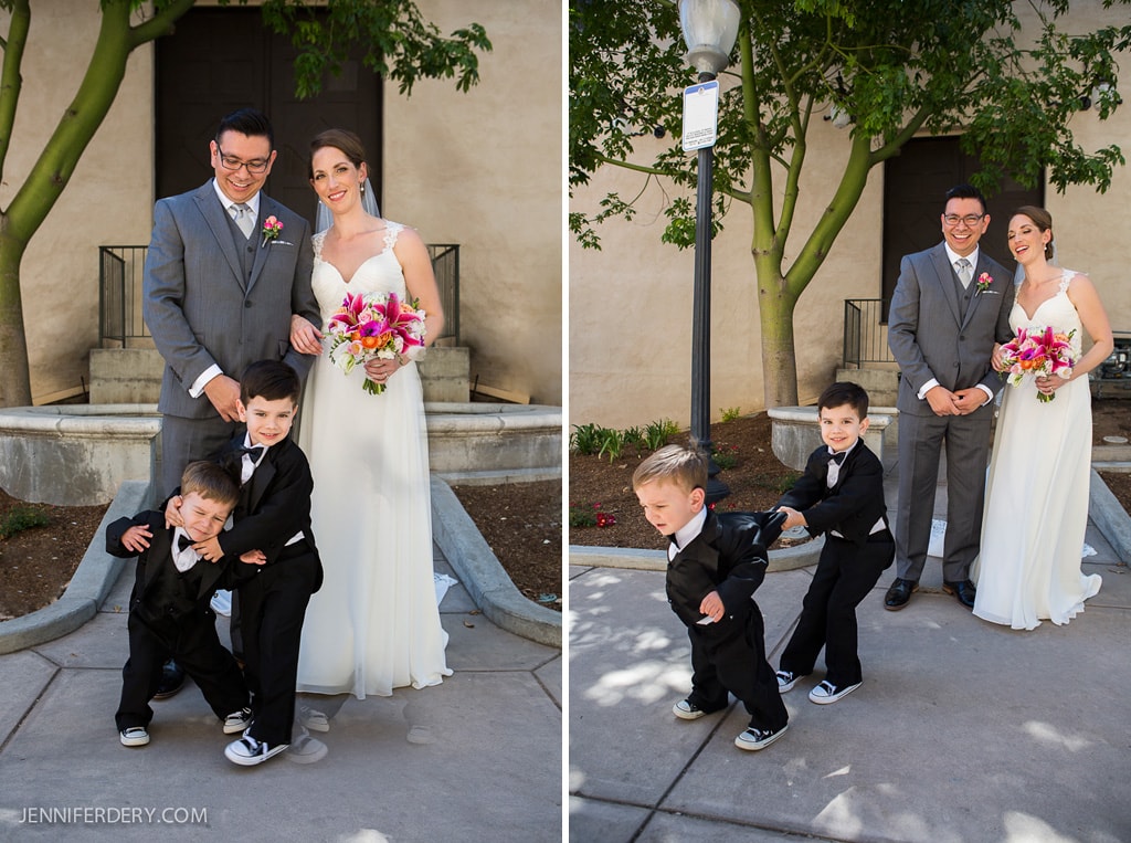A bride and groom pose for a photo, while two playful young boys in suits play in front of them. The boys are laughing and holding hands. The scene includes a building and trees in the background.