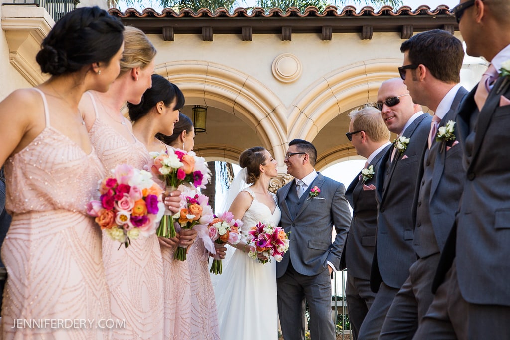 A bride and groom smile at each other surrounded by bridesmaids and groomsmen. The women wear pink dresses and hold colorful bouquets, while the men are in grey suits. They all stand before an arched building.