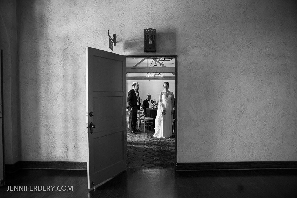 A black and white photo of a bride and groom standing in a room, viewed through an open door. They are accompanied by two seated individuals. The room has a patterned floor and a chandelier, with textured walls. A dark wooden floor is in the foreground.