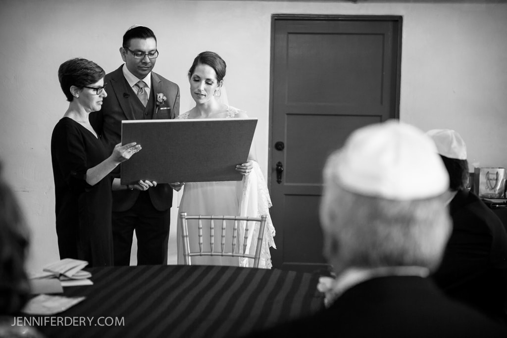 A black and white photo of a wedding ceremony. A couple, dressed formally, stands with an officiant holding a large document or board. The focus is on them, while seated guests, some wearing traditional caps, look on.