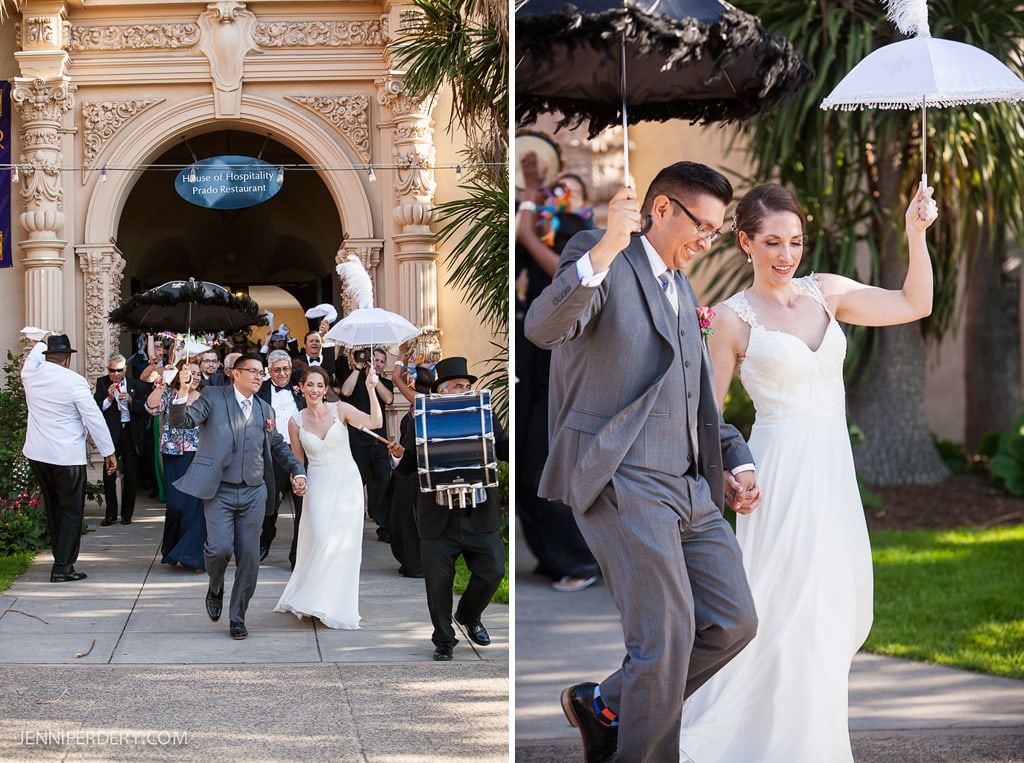 A joyful wedding procession with a bride and groom leading the way, holding parasols. The groom wears a gray suit, and the bride is in a white dress. Music plays as the group walks, with a band visible in the background under an ornate archway.