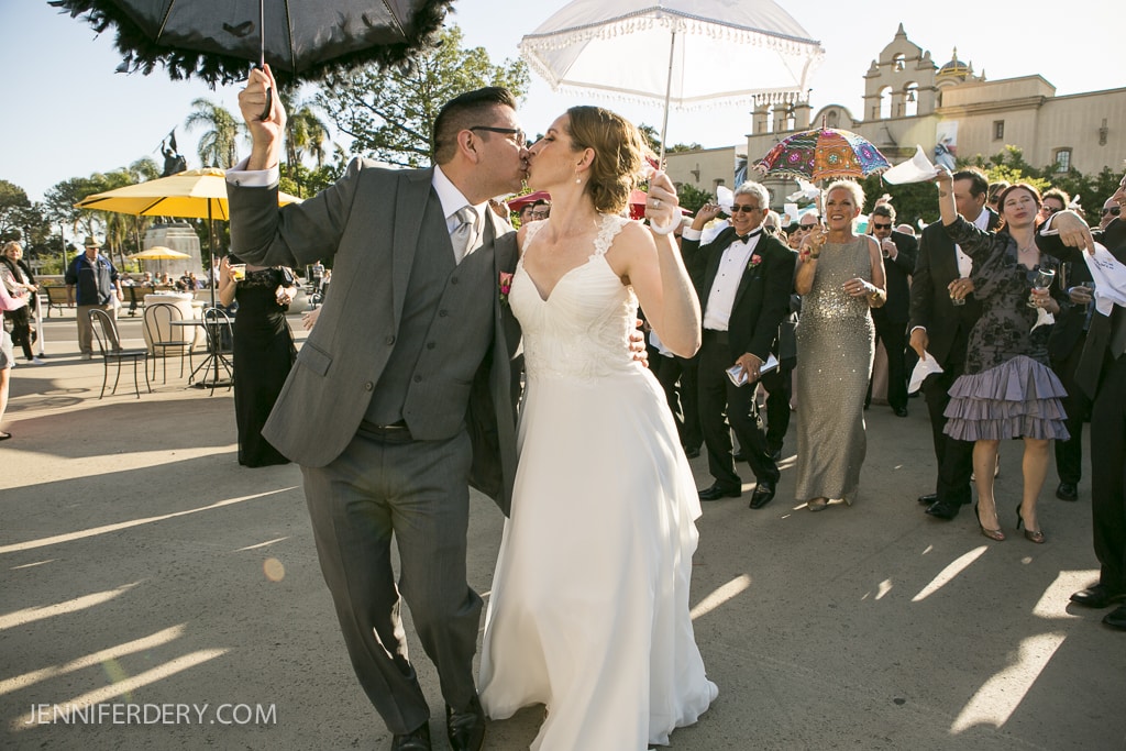A bride and groom kiss while walking with a lively wedding procession. The groom holds a black umbrella, and the bride holds white sunglasses. Guests carry colorful umbrellas and smile. Sunlit buildings and trees are in the background.