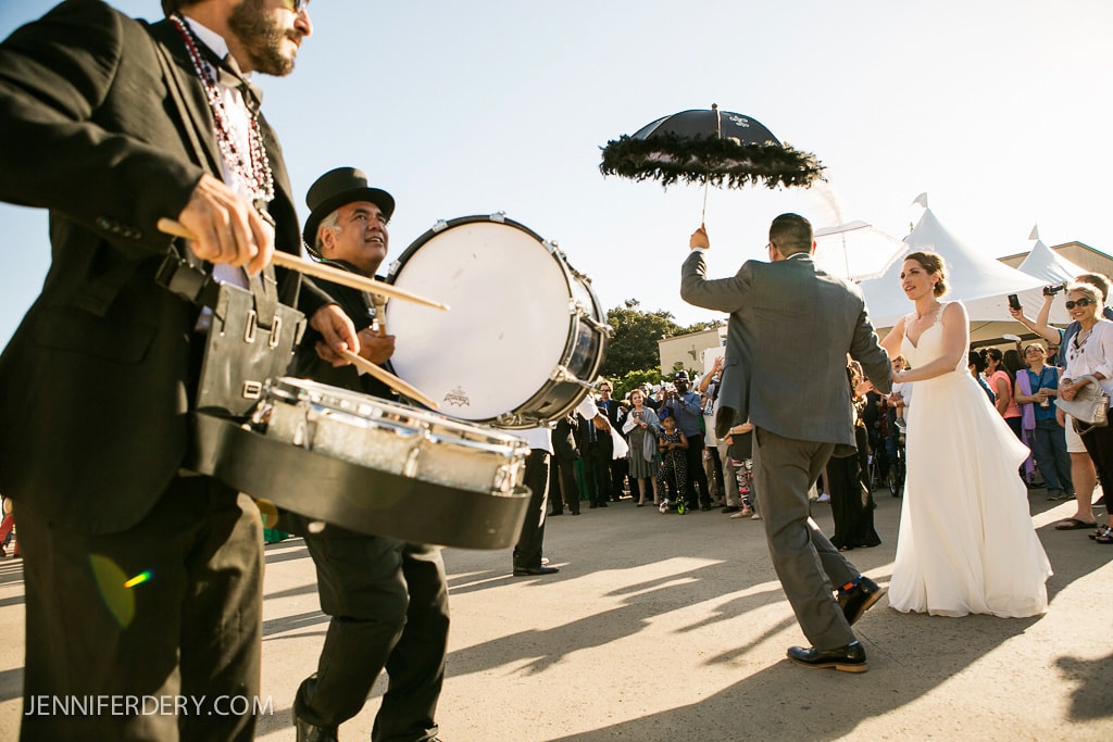A lively street procession featuring musicians in hats playing drums, a man in a suit holding a black parasol, and a woman in a white dress smiling, followed by a crowd of onlookers in a sunlit outdoor setting.