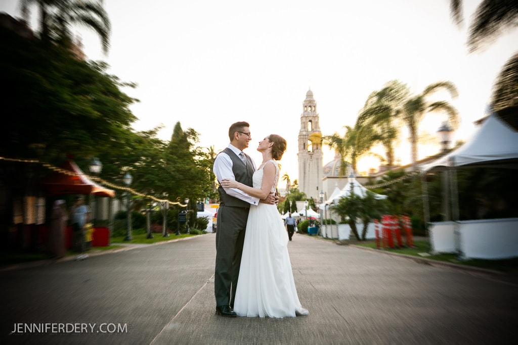 A couple in wedding attire shares an embrace on El Prado Way in Balboa Park lined with palm trees and tents. A historic tower is visible in the background, and the scene is bathed in soft, warm light.