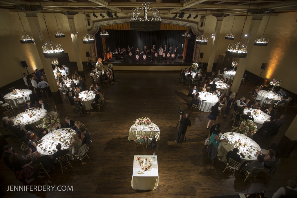 A spacious Prado ballroom with a wooden floor hosts a formal event. Several round tables with white tablecloths and floral centerpieces are arranged around the room. A band performs on a stage in the background. Guests are seated and mingling.