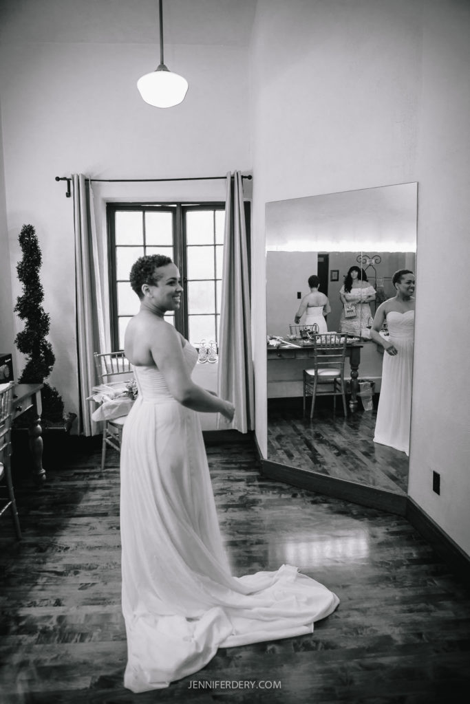 A bride in a strapless wedding dress stands in a room at Balboa Park, facing a large mirror. The room has wooden floors, a window with curtains, and a vanity in the background. Another person is reflected in the mirror, standing near the vanity. (Photo by Jennifer Dery).