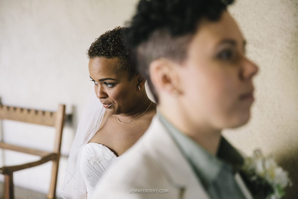 A person in a strapless white dress and veil stands behind another person in a white suit jacket and green shirt. They look affectionately towards their partner, who is in the foreground with a blurred face. They are indoors beside wooden chairs, capturing a beautiful moment at their Balboa Park wedding.