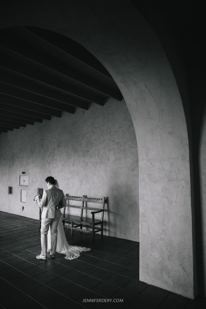 A black and white photo of a couple, dressed in wedding attire, standing close and holding hands under an archway. A wooden bench and some wall decor are visible behind them, capturing the rustic elegance of a Balboa Park wedding.