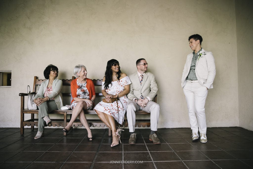 A group of five adults, including four sitting on a wooden bench and one standing at its end, are engaged in conversation. They are dressed in semi-formal attire, mostly in light colors. The scene resembles a Balboa Park wedding setting with a plain, light-colored wall and tile floor as the backdrop.