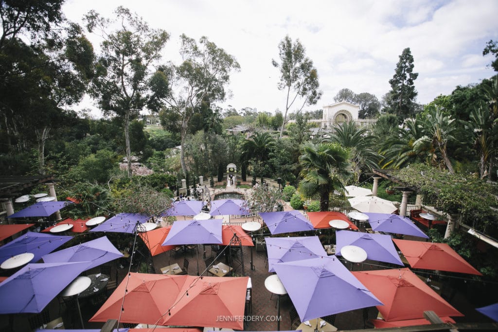 Aerial view of an outdoor seating area with tables and colorful umbrellas in shades of purple, blue, and red. The area is surrounded by tall trees, greenery, and a large building in the background under a partly cloudy sky—perfect for a Balboa Park wedding.