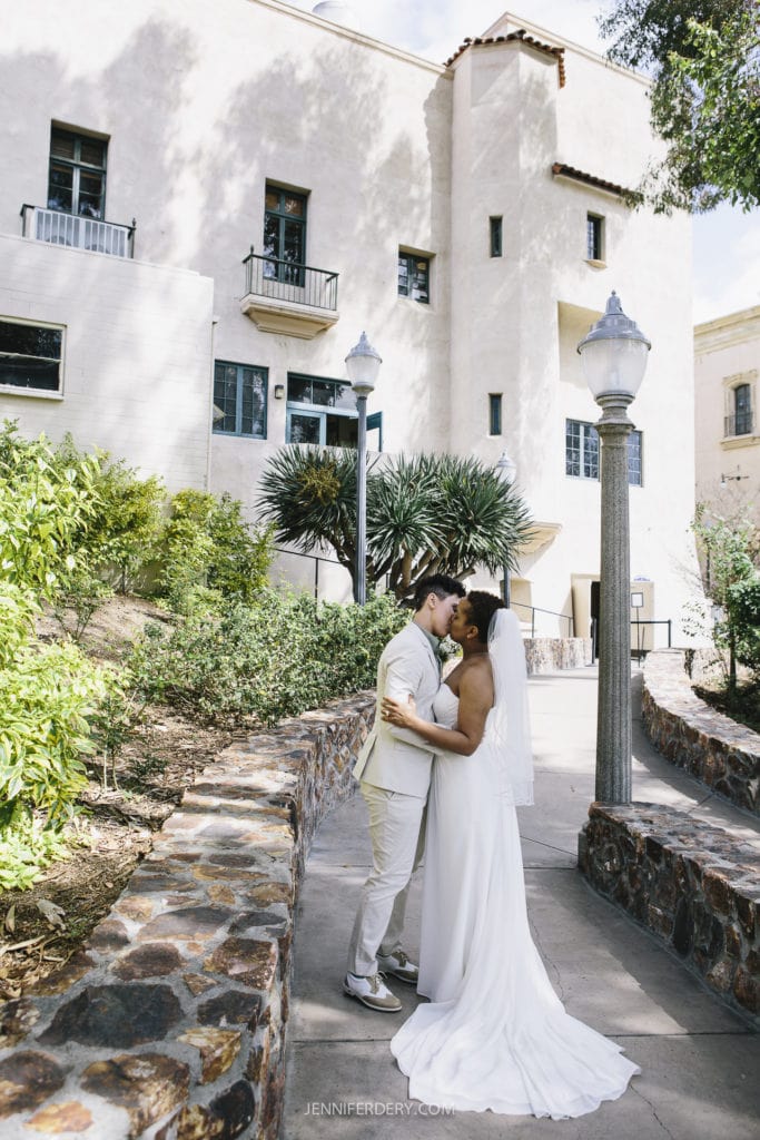 A newlywed couple shares a kiss on a path surrounded by greenery in Balboa Park, with a large cream-colored building in the background. The groom wears a light beige suit, and the bride dons a flowing white gown, holding a bouquet. Two vintage-style lampposts add charm to this picturesque wedding scene.
