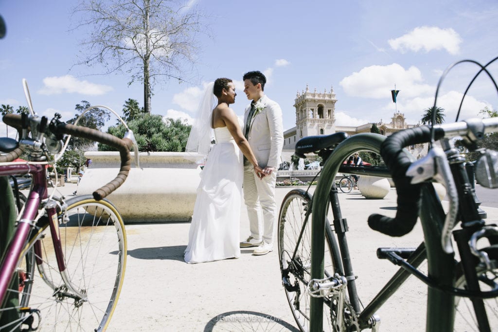 A bride and groom stand facing each other, holding hands and smiling, surrounded by bicycles in an outdoor setting. The sky is partly cloudy, and an ornate building can be seen in the background at their Balboa Park wedding.