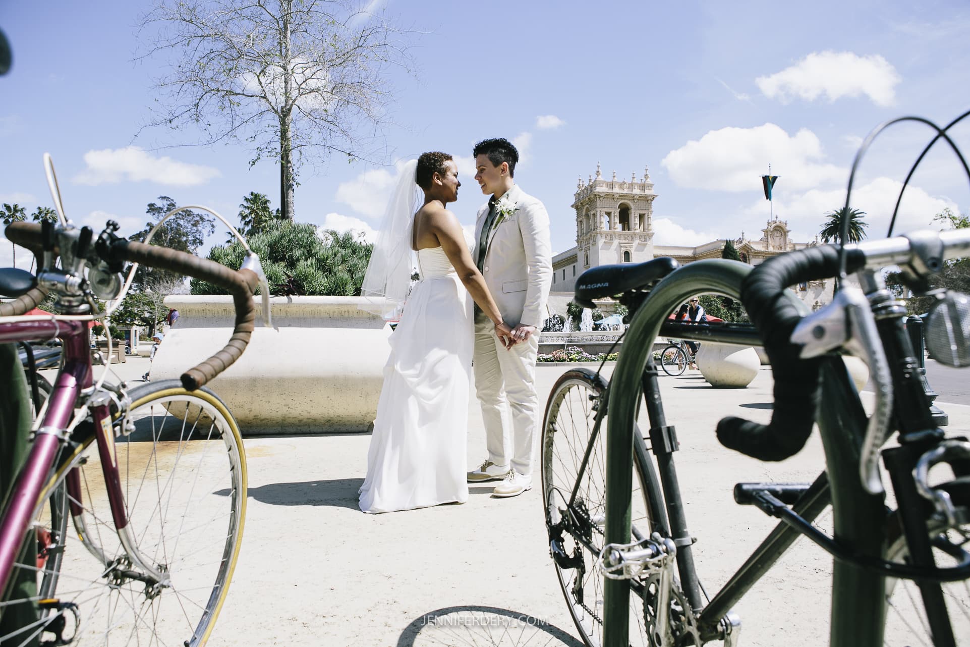 A bride and groom stand facing each other, holding hands and smiling, surrounded by bicycles in an outdoor setting. The sky is partly cloudy, and an ornate building can be seen in the background at their Balboa Park wedding.