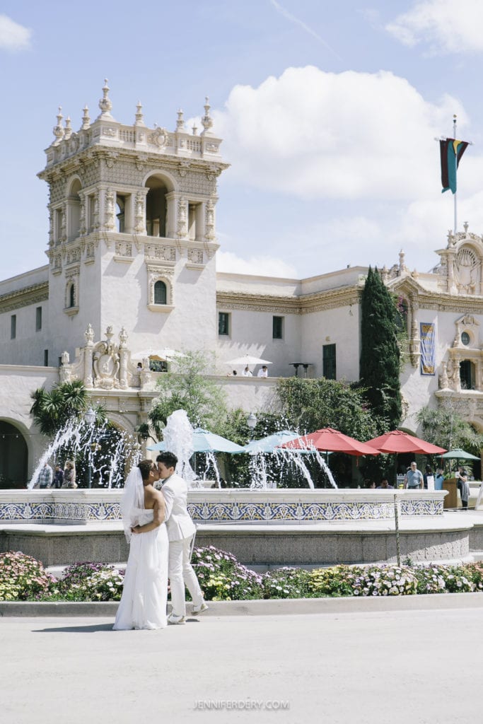 A couple dressed in white embraces in front of a fountain, with an ornate building showcasing detailed architecture in the background. Red umbrellas shield tables to the right, and blooming flowers line the fountain's edge. The sky is clear with a few clouds—a perfect day for a Balboa Park wedding.
