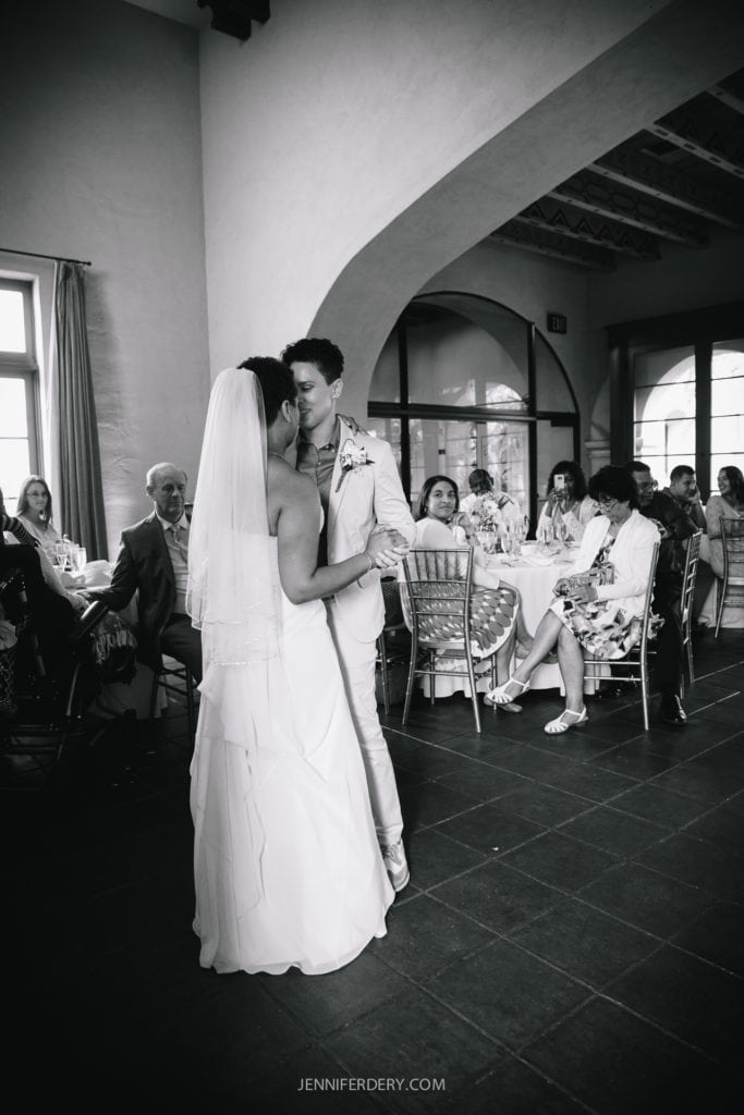 A black and white photo of two women dancing at their Balboa Park wedding. The bride on the left wears a long white dress and veil, while the other bride wears a white suit. Guests are seated at round tables in the background, watching the couple with smiles.