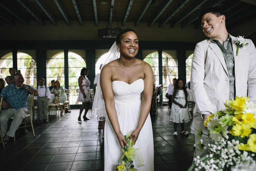 Two newlyweds in wedding attire smile joyfully at each other during their Balboa Park wedding ceremony. One wears a white strapless gown and veil, holding green leaves; the other wears a white suit with a floral boutonniere. Guests, including children, are in the background.