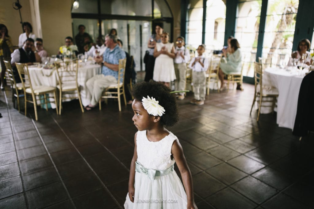 A young girl in a white dress with a flower headband stands in the foreground, looking away. In the background, guests are seated at a decorated banquet hall, some standing and clapping. The festive setting of their Balboa Park wedding boasts tables and chairs arranged neatly.