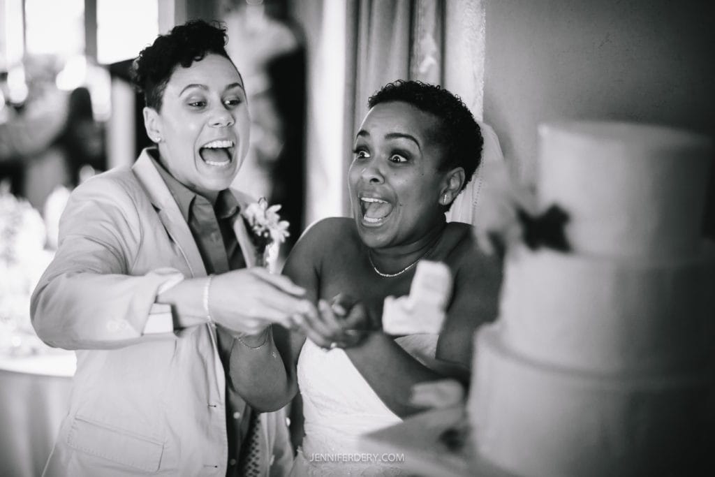 Two newlyweds are seen in a black and white image, both appearing joyful and surprised next to a multi-layered wedding cake. One is wearing a suit while the other is in a wedding dress, capturing an unexpected moment during their Balboa Park wedding celebration.