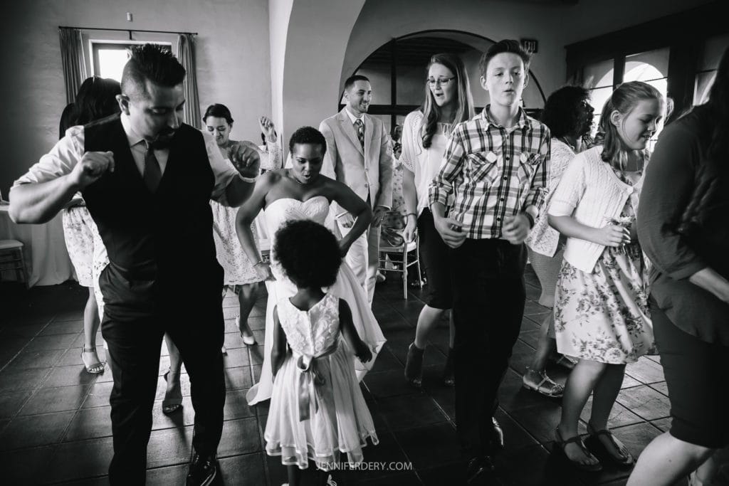 A group of people are dancing together indoors at a Balboa Park wedding, dressed in formal and semi-formal attire. The black and white image shows individuals, including children and adults, enjoying themselves, moving energetically with smiles and expressions of joy.