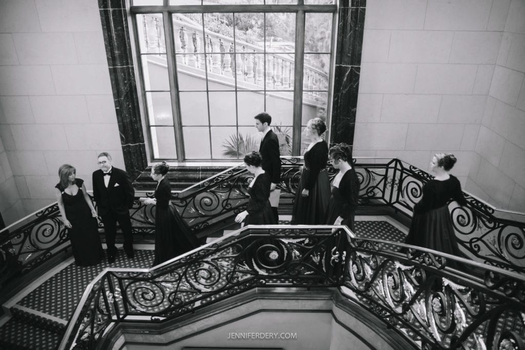 A black-and-white photograph of elegantly dressed people on an ornate grand staircase with intricate railing. A large window in the background illuminates the scene. Some individuals stand and chat while others descend the stairs.
