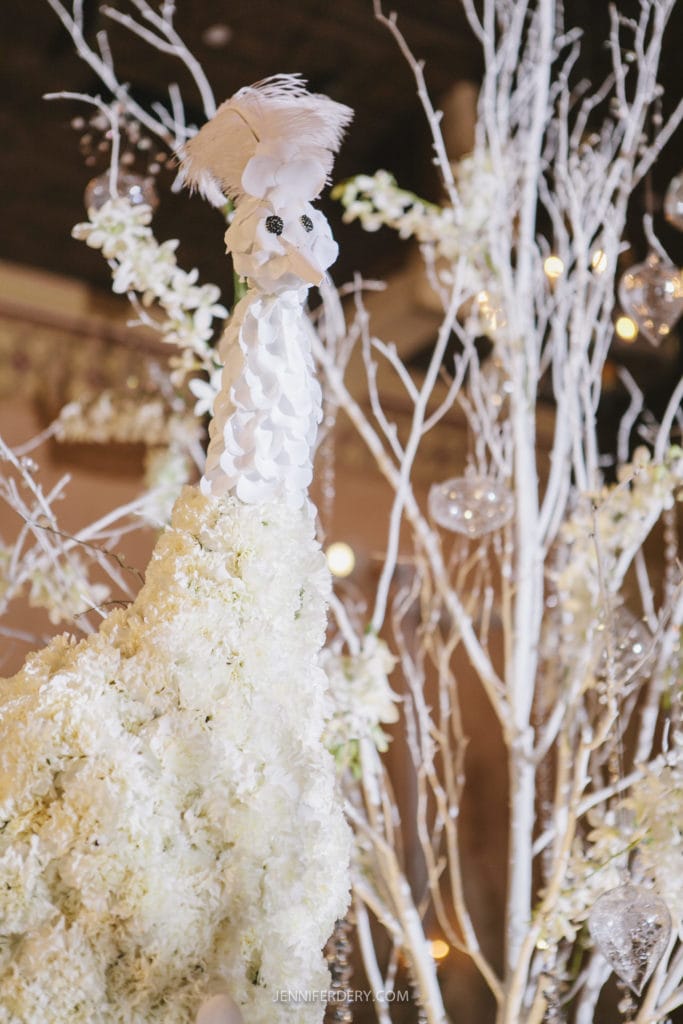 A sculpture of a peacock made entirely of white flowers stands against a backdrop of white tree branches adorned with hanging glass ornaments. The peacock has a feathery crest and detailed features, creating an elegant and artistic display.