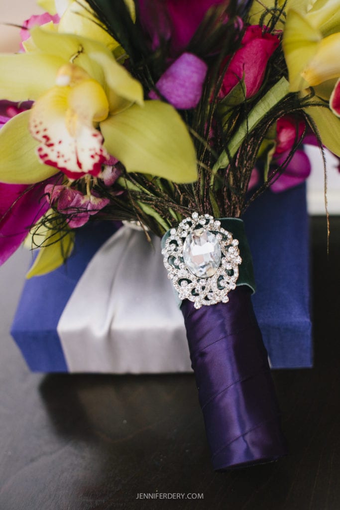 A bouquet featuring yellow and pink flowers, wrapped in a dark purple ribbon adorned with a sparkling jeweled brooch. The bouquet sits atop a blue gift box with a white ribbon, placed on a dark surface.