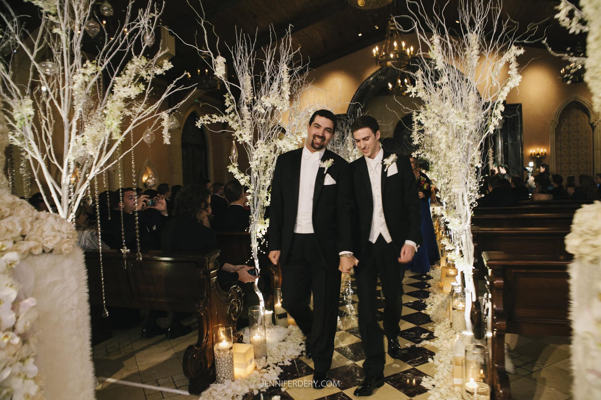 Two grooms in tuxedos walk hand-in-hand down an elegant, candle-lit aisle adorned with white flowers and decorative branches. They are smiling, surrounded by seated guests inside a dimly-lit, ornate venue, celebrating their wedding ceremony.