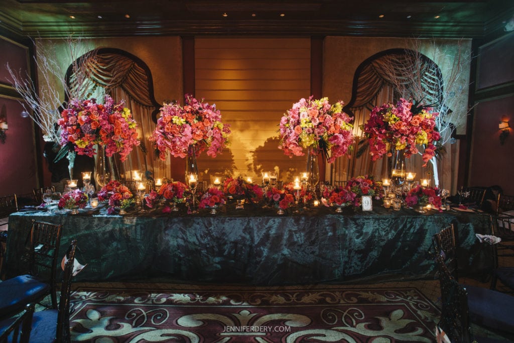 A lavishly decorated dining table with tall floral arrangements featuring pink, red, and yellow flowers. Lit candles are placed among the flowers, creating a warm, inviting atmosphere. The setting includes elegant drapery and dim lighting in a sophisticated room.