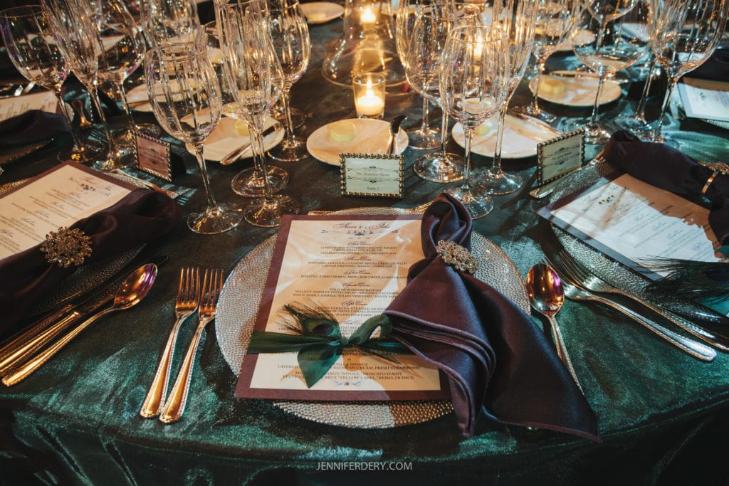 A lavishly set banquet table with luxurious decor. Each place setting includes a detailed menu, a dark napkin with an ornate brooch, multiple wine glasses, and sparkling silverware. A candle glows in the background, enhancing the elegant ambiance.