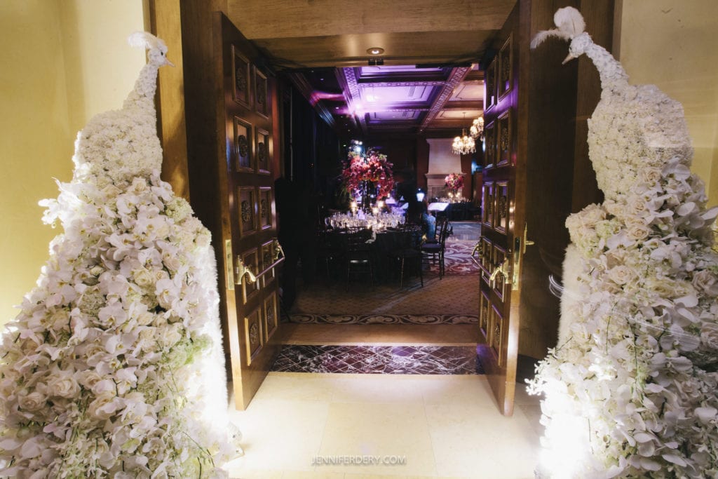 An opulent entrance decorated with two large, white floral peacock sculptures on either side, leading into a luxurious dining area adorned with elegant chandeliers, intricately designed ceiling, and tables set up with fine dining ware and red floral centerpieces.