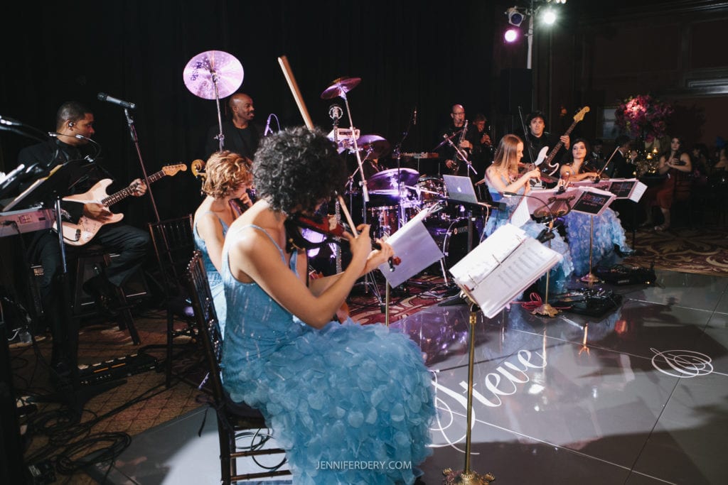 A live band performs on stage with several string musicians and a drummer. The musicians are dressed in matching blue outfits and play violins, cellos, and other instruments. The stage is well-lit, and the backdrop is dark. The atmosphere appears vibrant and lively.