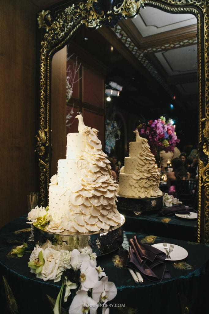 A large, ornate wedding cake with multiple tiers is adorned with intricate white frosting designs resembling feathers, and features a white peacock topper. The cake is placed on a reflective table with floral decorations, set against a lavish, gilded mirror.