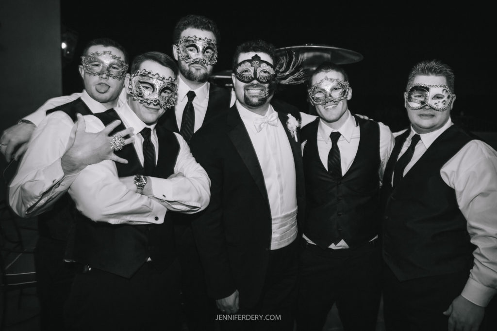 A group of six men in formal attire, all wearing decorative masquerade masks, stand closely together and smile at the camera. One man playfully sticks out his tongue. The image is in black and white.