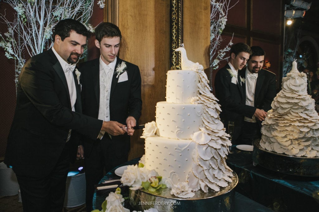 A wedding reception scene featuring two grooms in tuxedos cutting a large, multi-tiered white wedding cake adorned with intricate floral decorations. Both appear focused on the cutting task. Guests in the background also watch the cake-cutting ceremony.