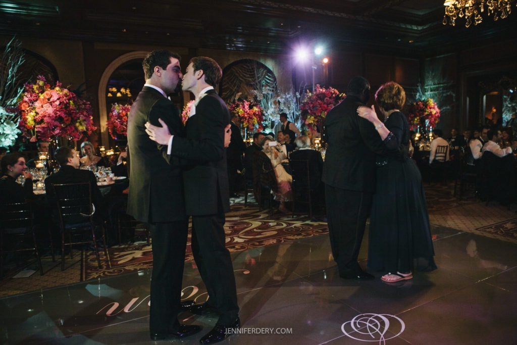 Two couples are dancing on a dimly lit dance floor at a formal event. One couple, two men in suits, share a kiss. The other couple, a man in a tuxedo and a woman in a long dress, dance nearby. The background features guests seated at tables adorned with tall floral arrangements.