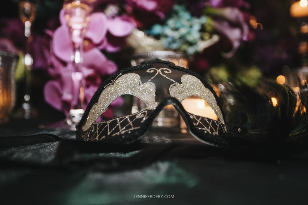 A close-up of a decorative masquerade mask resting on a surface. The mask features glittering gold and silver patterns on a dark base, with intricate detailing. The background includes blurred floral arrangements and lit candles. The artist’s website is noted at the bottom: jenniferdery.com.