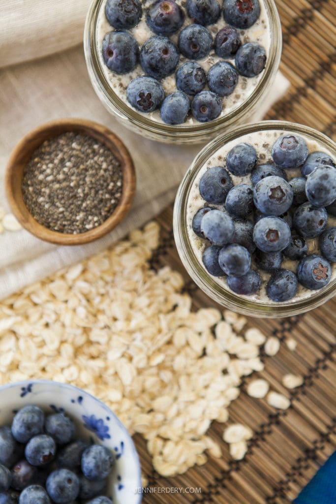 Top view of two jars filled with chia overnight oats topped with fresh blueberries. Surrounding the jars are rolled oats, a small bowl of fresh blueberries, and a bowl of chia seeds, set on a woven mat surface.