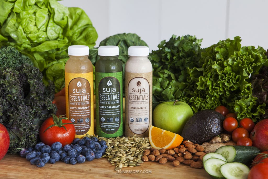 Three bottles of Suja Essentials juice (Spicy Lemonade, Green Greatness, Vanilla Nut) are placed on a wooden surface surrounded by fresh fruits, vegetables, seeds, and nuts, including apples, blueberries, tomatoes, cucumbers, lettuce, kale, and almonds.