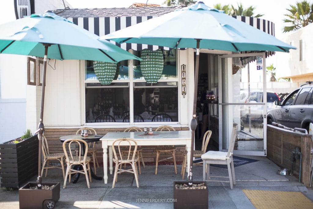 A cozy outdoor seating area of Little Lion Cafe with a white facade. Light wooden chairs and a white table are shaded by teal umbrellas. The interior of the cafe is partially visible through large windows adorned with green patterned decor.