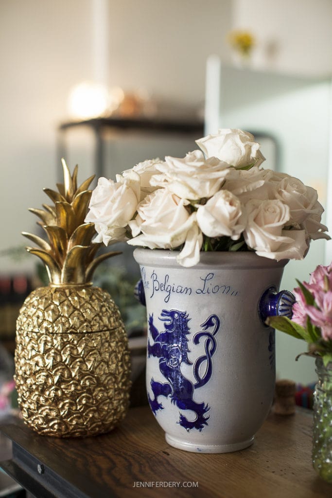 A decorative table holds a ceramic vase adorned with an intricate blue lion design filled with white roses. Next to it stands a golden pineapple-shaped sculpture. The vase has the words "The Belgian Lion" written on it. The background is blurred.