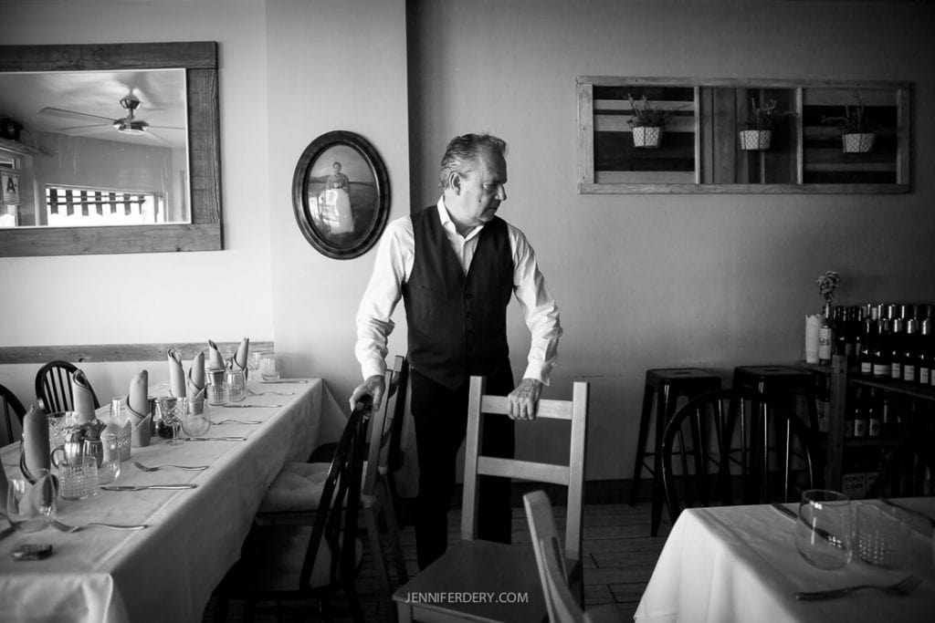 A person standing in the dining area at Little Lion Cafe, holding the back of a chair, appears to be organizing or setting up. Tables are set with tableware, and framed mirrors and plant decorations hang on the walls. The black-and-white image gives a classic, timeless feel to the scene.