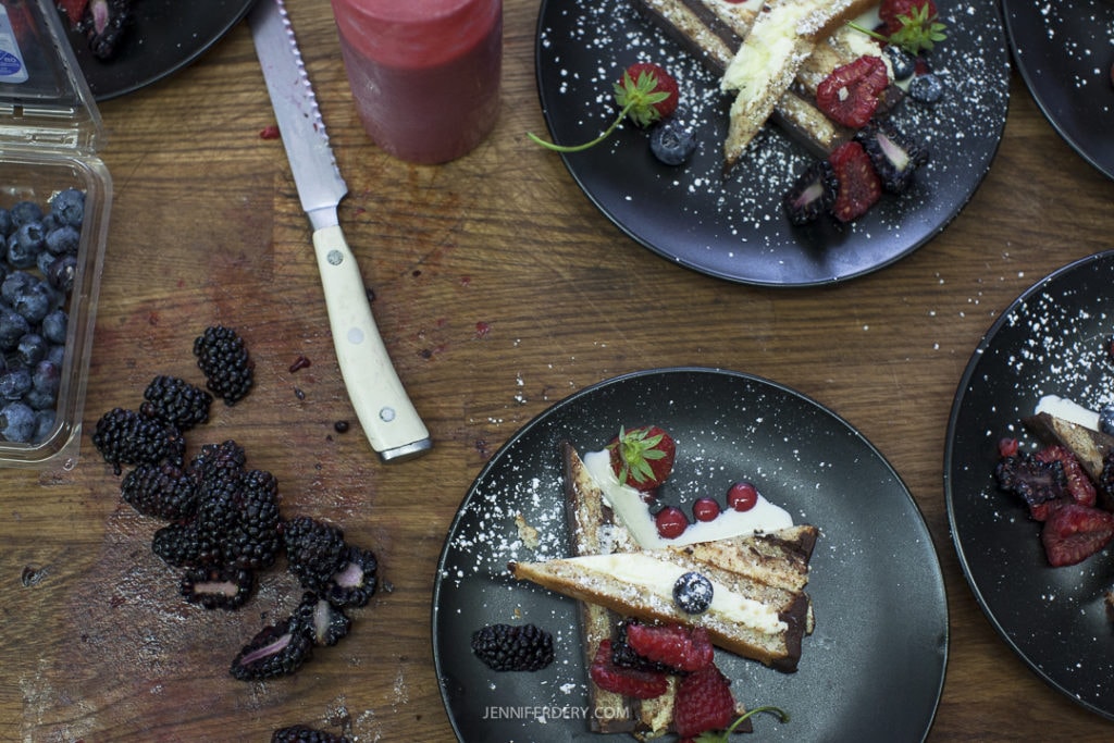 A wooden table is set with black plates holding slices of cheesecake topped with assorted berries. A knife lies on the table near a container of blueberries and a scattered pile of blackberries. A red candle is placed beside the dessert plates.