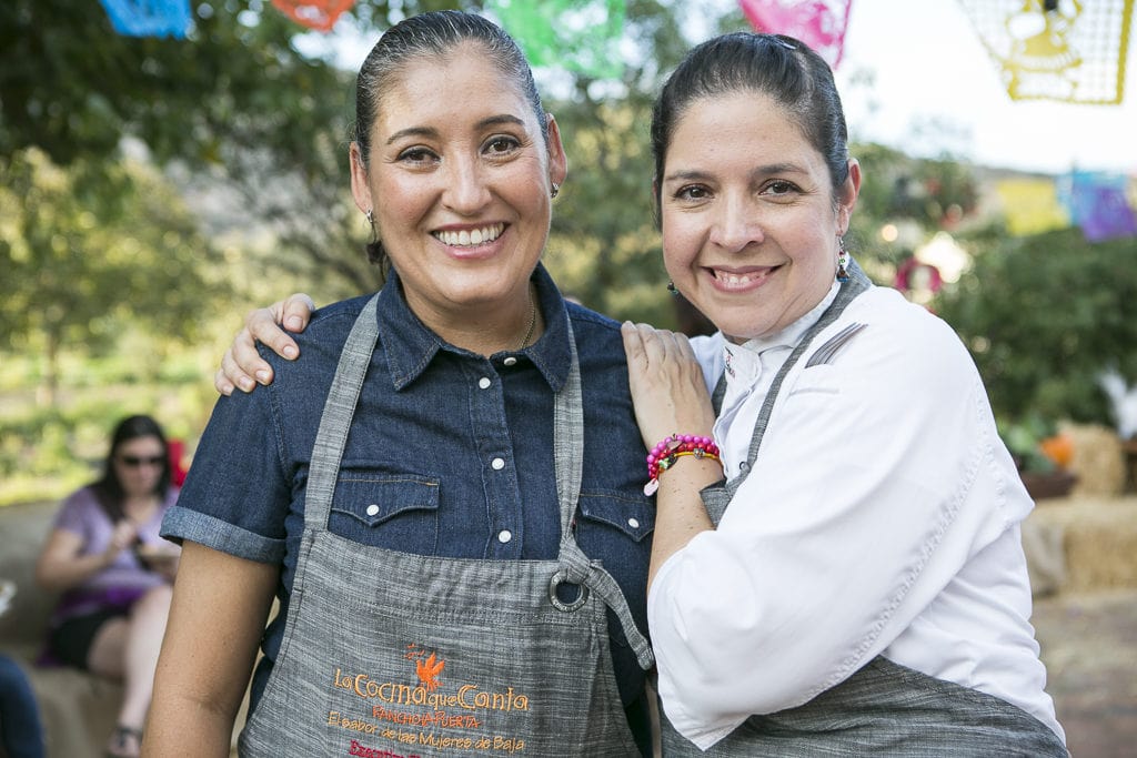 Two women smile warmly at the camera, one in a navy denim shirt and the other in a white top. Both wear aprons that say "La Cocina Que Canta." They are outdoors, with colorful paper decorations and trees in the background. Other people are blurred in the distance.