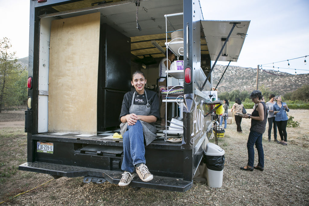 A smiling chef wearing an apron sits on the edge of an open food truck. Behind them, various kitchen utensils are visible inside the truck. In the background, people are seen talking and enjoying the outdoor setting at Rancho La Puerta Spa with mountains and trees.