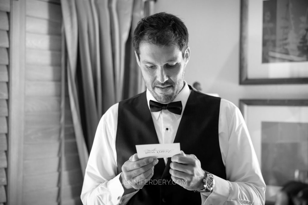 A man dressed in a tuxedo, wearing a bow tie and a vest, reads a small piece of paper. The background features curtains and framed pictures on the walls. The atmosphere appears formal, suggesting a special event or occasion.