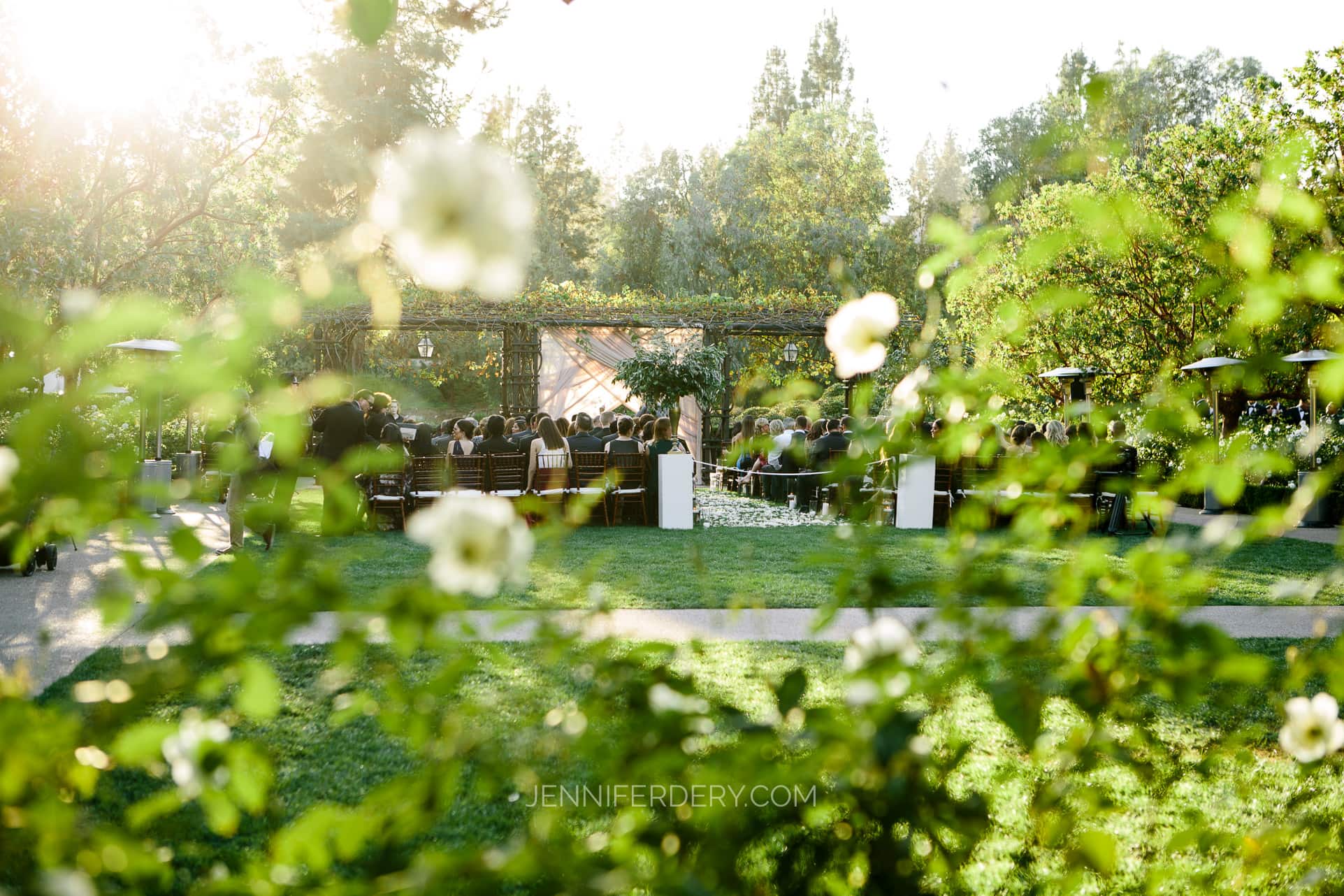 A sunlit outdoor wedding ceremony at Rancho Bernardo Inn with guests seated in rows, viewed through greenery and white flowers. A floral arch and trees are visible in the background, creating a serene, natural setting. For those wondering how to inquire about wedding photography for such moments, capturing the essence is key.